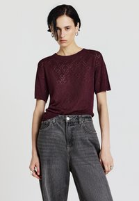 Maglione lavorato a maglia bordeaux con maniche corte, caratterizzato da un motivo a diamante. Abbinato a jeans neri sbiaditi a vita alta.