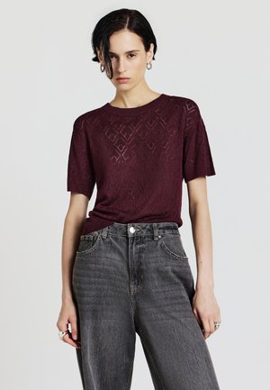 Maglione lavorato a maglia bordeaux con maniche corte, caratterizzato da un motivo a diamante. Abbinato a jeans neri sbiaditi a vita alta.