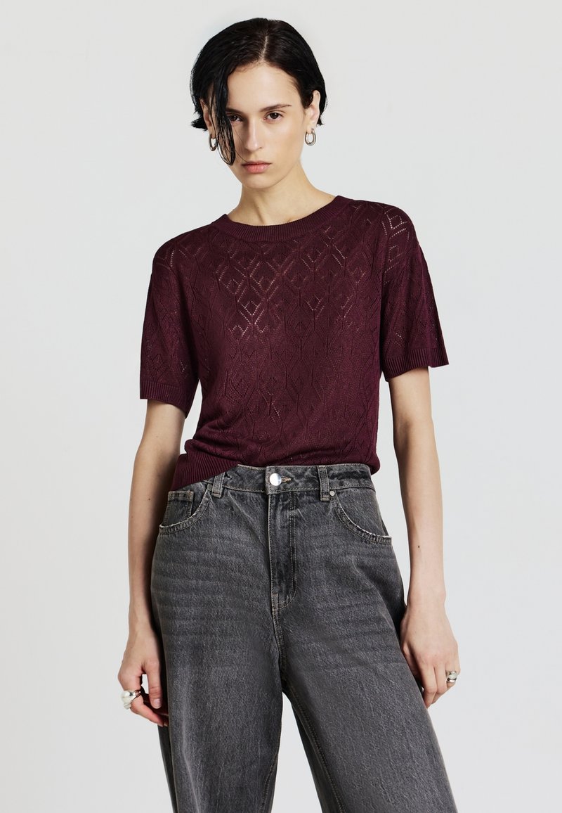 Maglione lavorato a maglia bordeaux con maniche corte, caratterizzato da un motivo a diamante. Abbinato a jeans neri sbiaditi a vita alta.