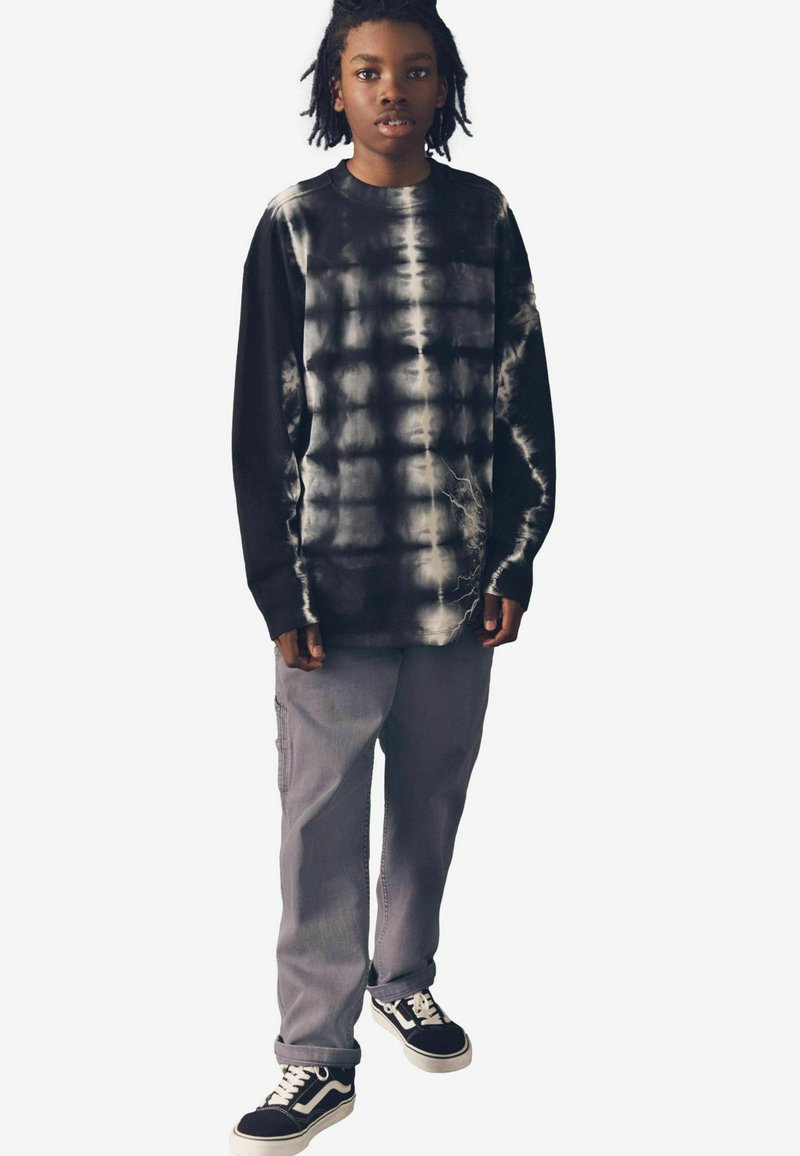 Langärmliges schwarzes Batikshirt mit weißen Mustern, kombiniert mit grauen Cargo-Hosen und schwarzen Sneakers mit weißen Schnürsenkeln und Akzenten.
