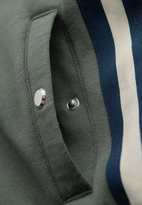 Tissu vert avec une texture, comportant une poche latérale, un bouton-pression argenté et des accents rayés bleu marine et crème. Vue en gros plan.