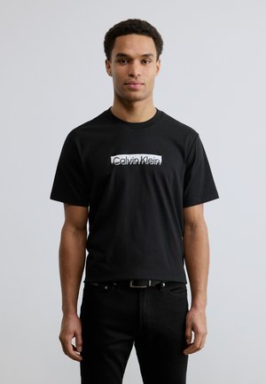 Czarny bawełniany T-shirt z okrągłym dekoltem, z białym logo „Calvin Klein” na piersi, noszony z czarnymi dżinsami.