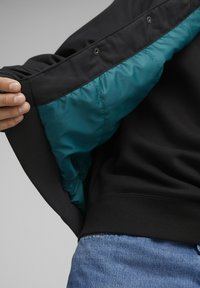 Svart jakke med tealfarga fòr, med trykknapper langs halsen og ribbestrikka mansjetter. Myk, glatt tekstur, uformelt design.