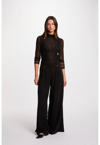 Vrouw met lang, donker haar, gekleed in een zwarte doorschijnende top met geometrisch patroon, hoge taille zwarte broeken met wijde pijpen en zwarte schoenen.