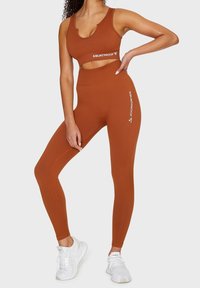Femme portant un soutien-gorge de sport côtelé couleur rouille et des leggings avec le texte "SQUATPROOF", posant avec une main sur la hanche, portant des baskets blanches Adidas.
