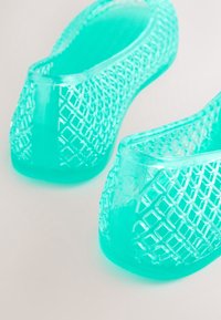 Chaussures en gelée turquoise translucide avec un motif en treillis, montrant un gros plan du talon et des détails latéraux sur un fond clair.