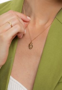 Auksinė pakabuko grandinėlė su apvaliu dizainu ir kryžiaus detale, kabančia ant subtilios grandinėlės, dėvima ant nuogos odos ir žalios striukės.