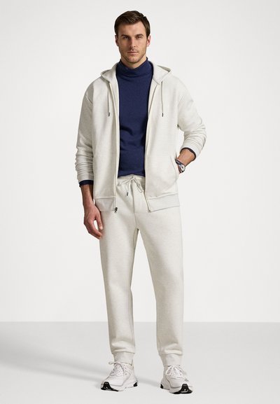 Light grey zip-up hoodie with drawstrings, paired with matching joggers. Underneath, a navy turtleneck. White sneakers complete the outfit.