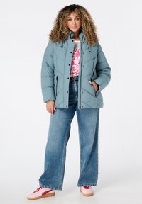 Gevoerde teal jack met een hoge kraag, ruitvormig gestikt ontwerp en met rits afsluitbare zijzakken, gecombineerd met wijde jeans en roze sneakers.