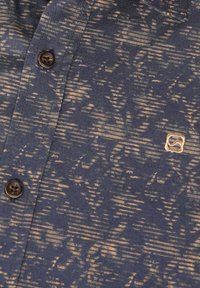 Camisa de botones en tela azul marino con un sutil patrón beige. Presenta un pequeño logo bordado y botones marrones. Textura suave.