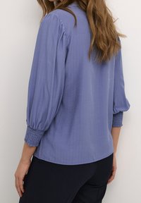 Blouse bleue texturée avec des manches bouffantes et des poignets froncés. Présente un ourlet droit et un délicat motif en rayures verticales.