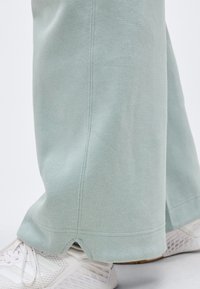 Φαρδιά παντελόνια ανοιχτού πράσινου με μικρό σκίσιμο πάνω από το τελείωμα, φορεμένα με λευκά αθλητικά παπούτσια από δίχτυ, σε λευκή επιφάνεια.