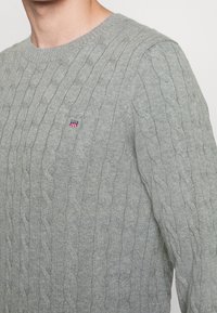 Šedý svetr s kulatým výstřihem a vzorem kabelového pletení. Malý emblém na prsou dodává kontrastní detail.