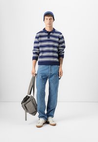 Gestreepte marineblauwe en witte gebreide trui met een kraag, gecombineerd met lichtblauwe jeans. Houdt een grijze schoudertas vast, draagt witte sneakers en een beanie.