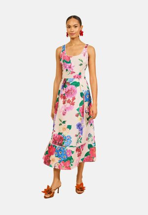 Kobieta ubrana w sukienkę bez rękawów do kostek w kwiatowy wzór w kolorach różowym, czerwonym i niebieskim, czerwone kolczyki w kształcie kwiatów oraz pomarańczowe sandały na obcasie z detalem kwiatowym.