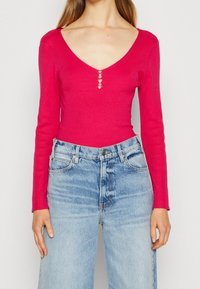 Frau trägt ein enges, knallpinkes, geripptes Langarmoberteil mit drei kleinen Knöpfen am Ausschnitt und hellblauen, hochgeschnittenen Jeans.