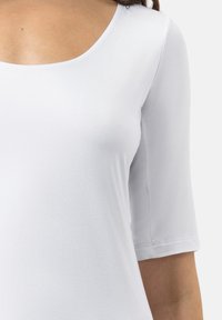 Vrouw draagt een eenvoudige witte nauwsluitende top met een ronde hals en ellebooglange mouwen tegen een lichte achtergrond.