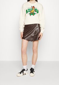 Gräddvit sweatshirt med grön och svart grafisk tryck, kombinerad med en brun läder mini-skjorta med ett diagonalt omslag. Svart och vit sneakers.