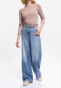 Frau trägt ein beiges langärmliges Oberteil, hellblaue weit geschnittene Jeans mit brauem Gürtel und beige Sneakers mit Leopardenmuster-Details.
