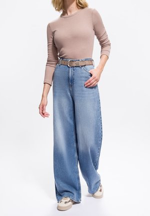 Femme portant un haut beige à manches longues, un jean bleu clair à jambes larges avec une ceinture marron, et des baskets beiges avec des détails imprimés léopard.