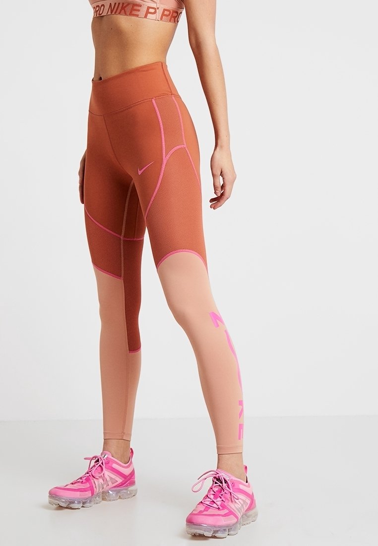 Legginsy sportowe Nike z wysokim stanem, siateczkowymi i gładkimi panelami w kolorze rdzy i beżu, z różowymi akcentami oraz logo Nike. W zestawie z różowymi sneakersami.
