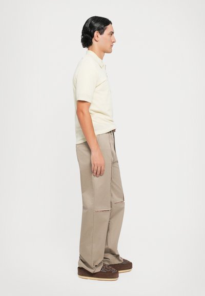 Lichtgele polo met korte mouwen, gecombineerd met beige broeken met zijsplitten en bruine instap-schoenen met geborduurde details.