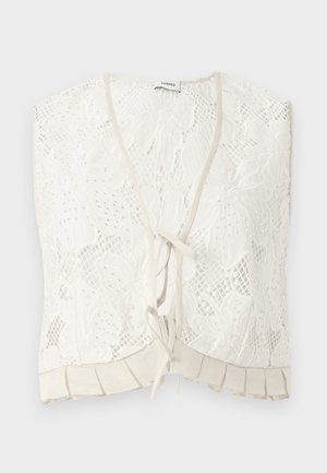 Kvinde iført en beige floral blonde crop top med flæsekant og matchende lang blonder nederdel, kombineret med sorte Mary Jane-sko.