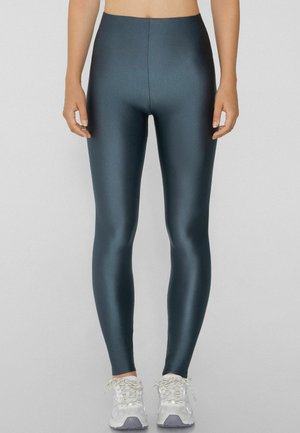 Leggings de cintura alta en un material brillante de color verde azulado oscuro, con un diseño ajustado y textura suave, combinados con zapatillas blancas.