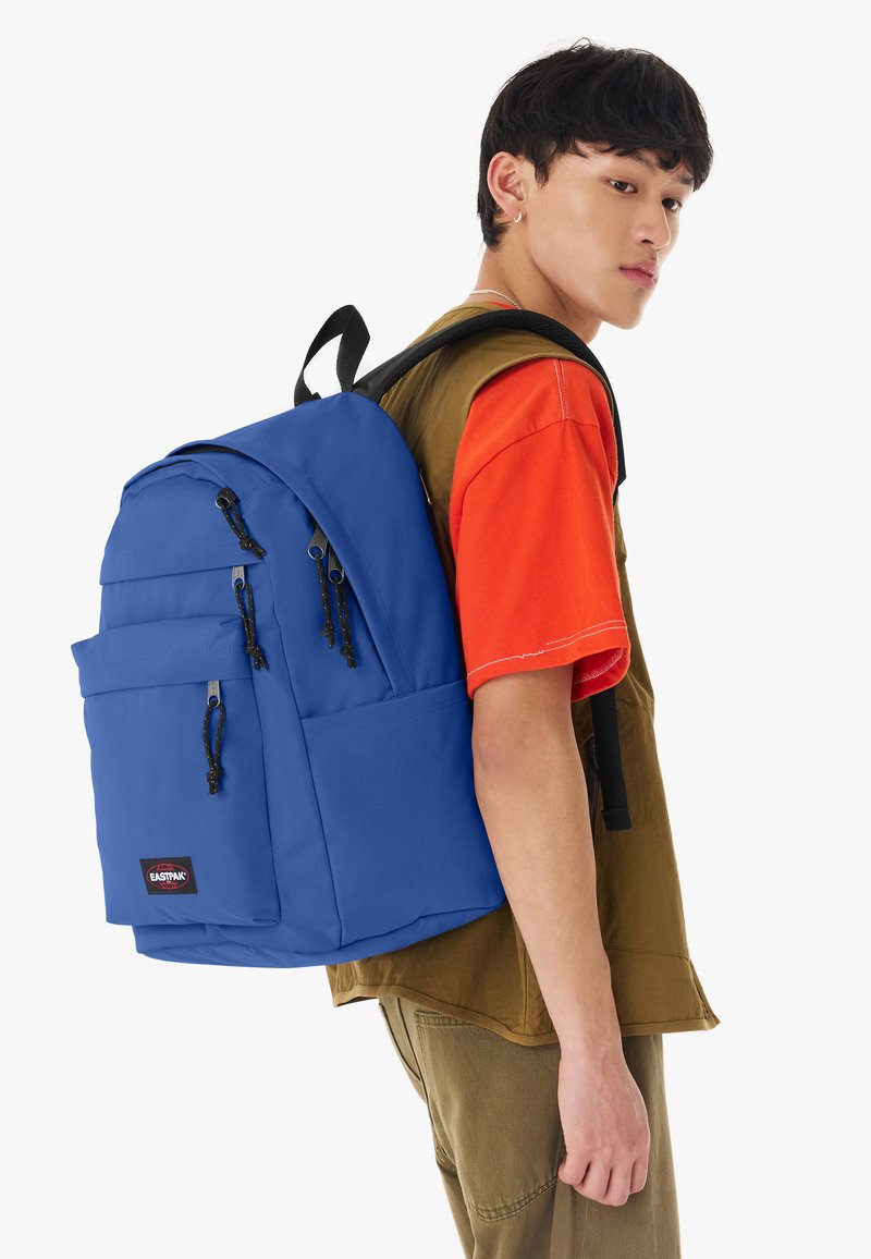 Jeune homme portant une chemise orange et un gilet marron porte un grand sac à dos bleu avec plusieurs poches zippées dans son dos.