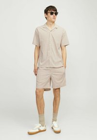Chemise à manches courtes, boutonnée, et short assorti en crème avec un motif à carreaux marron, porté avec des baskets blanches et des chaussettes grises.