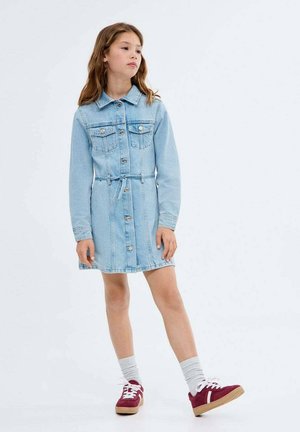 Ragazzina in piedi con un vestito di jeans blu chiaro a bottoni, maniche lunghe, vita annodata, calze bianche e scarpe da ginnastica rosse con suole bianche.