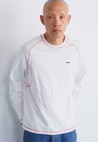 White long-sleeve shirt with red stitching accents and a small black "OBEY" logo on the chest. Fabric is smooth cotton.
