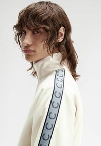 Sudadera de color crema, con cuello alto y franjas laterales grises, que presenta un patrón de emblemas circulares. Textura suave y diseño ajustado.