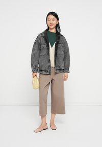 Jovem mulher com uma jaqueta oversized de denim preta, camisa verde, cardigan bege, calças largas e curtas, segurando uma pequena bolsa amarela, usando sapatilhas cor-de-rosa.
