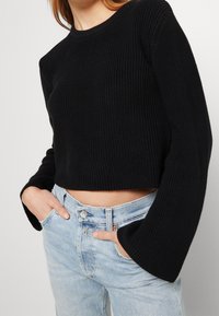 Schwarzer, gerippter, kurz geschnittener Pullover mit weiten Ärmeln, getragen über hellblauen, hoch taillierten Jeans. Strukturierter Stoff und Rundhalsausschnitt sichtbar.