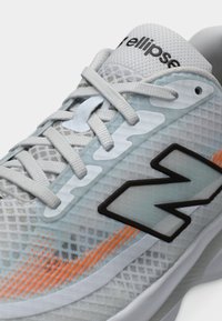 Άσπρο και γκρι αθλητικό παπούτσι με πλέγμα, λευκά κορδόνια, πορτοκαλί σχέδιο κοντά στη σόλα και μεγάλο λογότυπο «N» με μαύρο περίγραμμα στο πλάι.