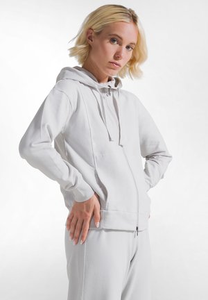 Blondynka ubrana w jasnoszarą bluzę z zamkiem i dopasowane spodnie, stojąca z rękami na biodrach na jednolitym tle.