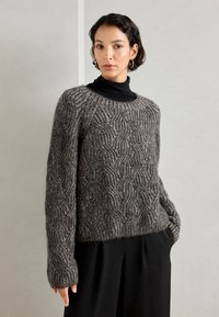 Sweter dzianinowy w kolorze szarym z fakturowanymi wzorami w fale, z długimi rękawami i luźnym krojem. Nosi się go na czarnym golfie, co podkreśla przytulny design.