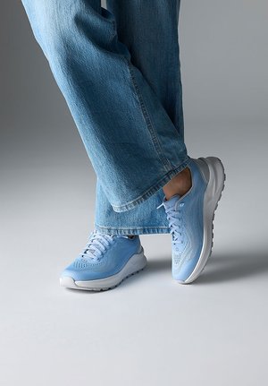 Person mit hellblauen Turnschuhen und weit geschnittenen blauen Jeans, die mit einem Fuß vor dem anderen auf einem grauen Boden steht.