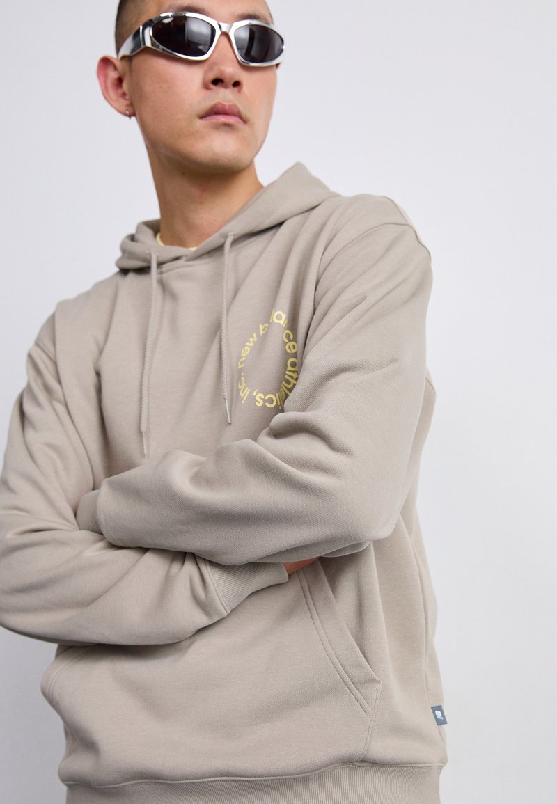 Haut à capuche beige en tissu doux, doté d'une poche kangourou et d'un logo circulaire doré sur la poitrine. Des lunettes de soleil argentées complètent le look.