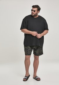 Fekete, túlságosan bő póló, camouflage pálma mintás rövidnadrág, fekete papucs fehér betűzéssel, kerek napszemüveg, sima háttér előtt áll.
