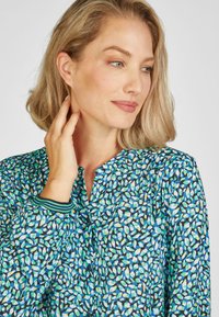 Blaue gemusterte Bluse mit grünen und weißen abstrakten Formen. Verfügt über einen runden Ausschnitt und Falten in der Nähe der Schultern. Texturierter Stoff.