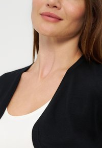 Unteres Gesicht und Hals einer Frau, die eine schwarze, vorne offene Strickjacke über einem weißen Oberteil trägt, mit langen braunen Haaren, die auf den Schultern sichtbar sind.