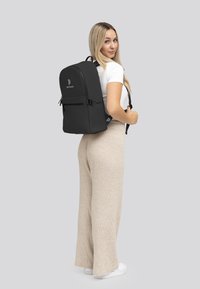 Schwarzer Rucksack mit einem glatten, strukturierten Design und Logo auf der Vorderseite, kombiniert mit einer Person in hellfarbiger Kleidung und weiten Hosen.