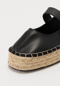 Chaussure en cuir noir avec un bout rond, présentant une plateforme en corde de jute et une sangle noire. La semelle est cousue avec un fil de contraste.
