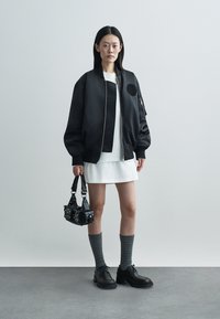 Giacca bomber nera con texture liscia, vestito bianco, calze grigie al ginocchio, scarpe nere e una piccola borsa nera con borchie.