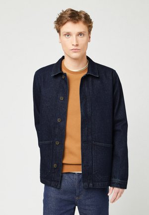 Donkerblauwe denimjas met een knoopsluiting aan de voorkant, twee zakken en een klassieke kraag. Draag dit over een bruine crewneck trui.