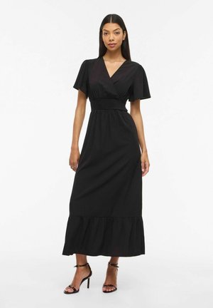 Kvinne stående i en svart midi-kjole med V-hals, korte flæseskjermmermer og høye hæler med ankelstropp på en ensfarget hvit bakgrunn.