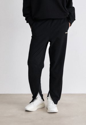 Persona con pantalones de chándal negros con aberturas laterales en los tobillos y zapatillas blancas, de pie sobre un suelo claro frente a una pared lisa.