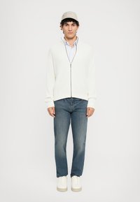 Bijela dukserica s patentnim zatvaračem i rebrastom teksturom, svijetloplava košulja s kragnom ispod, plave traperice i bijele tenisice. Sivi šešir zaokružuje izgled.